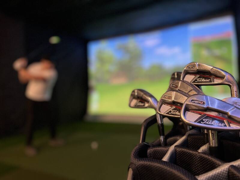 Golf kassi täynnä erilaisia mailoja ja taustalla golffaaja juuri lyömässä.