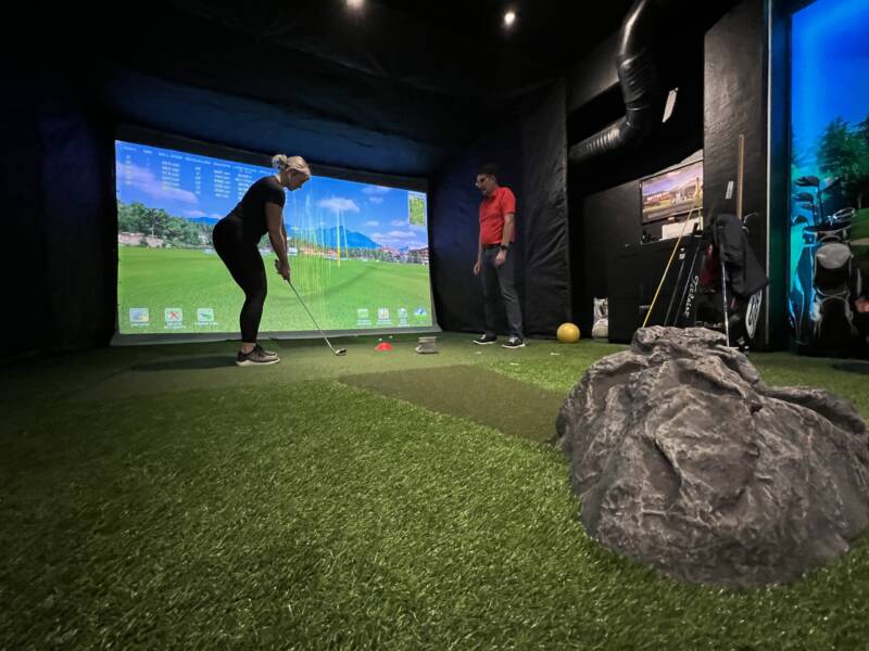 Pelaaja valmistautuu lyömään toisen varmistaessa asentoa golf simulaattorissa.